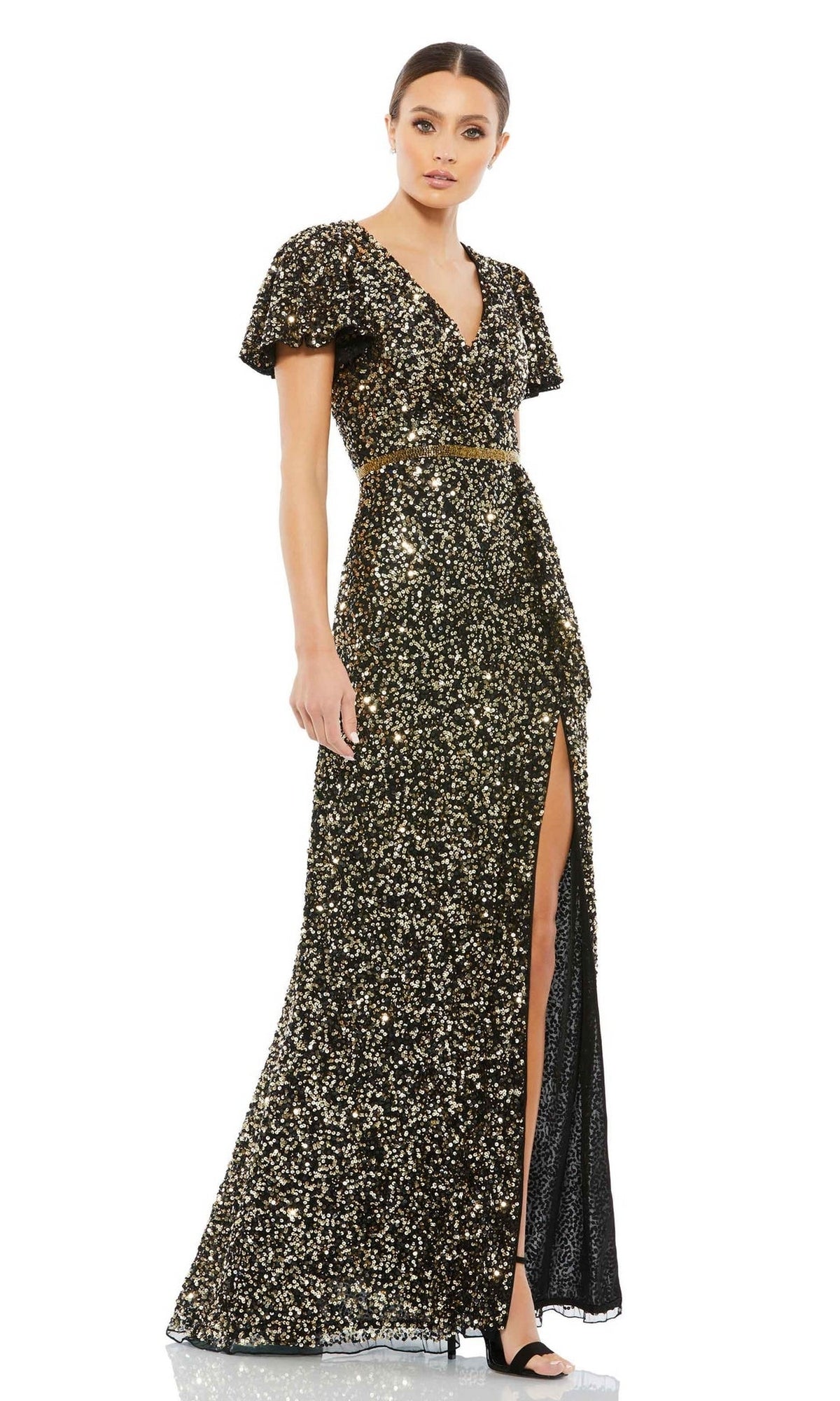 A woman wears the Mac Duggal 5540 Short-Sleeve Sequin Formal Dress—a black, floor-length gown with gold sequins, v-neckline, belted waist, high slit—paired with black heels.